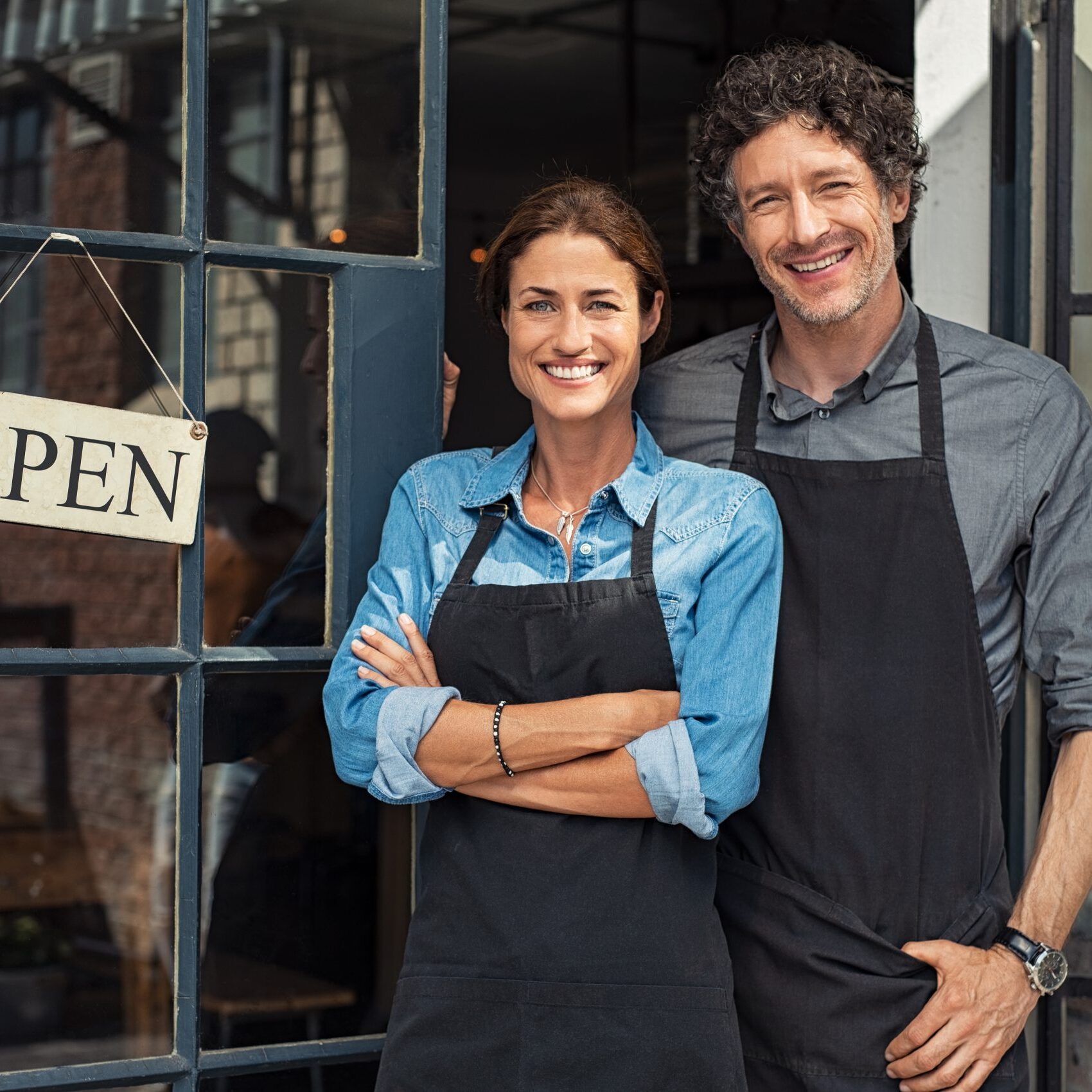Small business owners couple Two cheerful small business owners smiling and looking at camera while standing at entrance door. Happy mature man and mid woman at entrance of newly opened restaurant with open sign board. Smiling couple welcoming customers to small business shop.